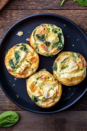 Overhead view of four Turkey and Brie Egg Bites with spinach, turkey, melted Brie, and herbs on a matte black plate.