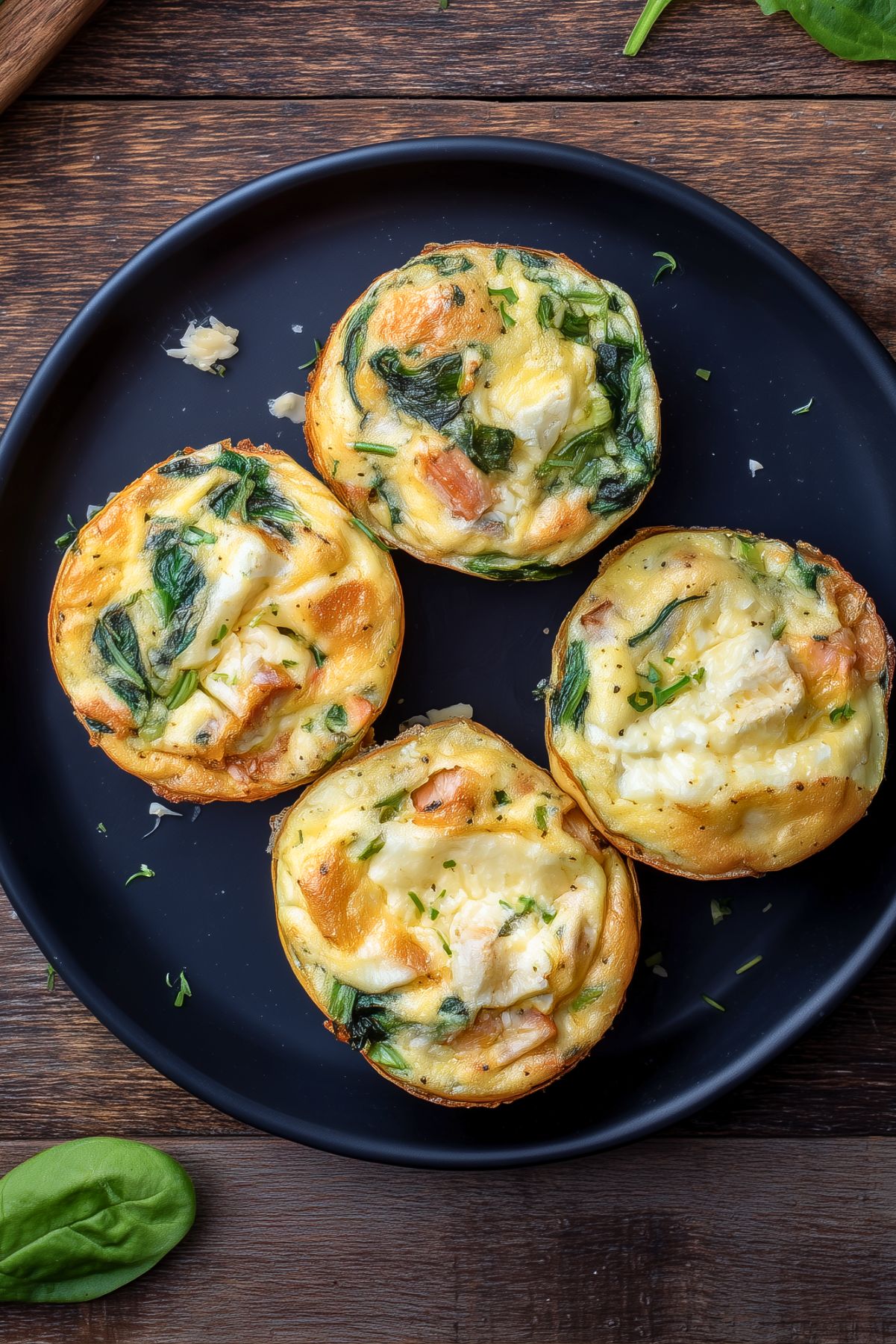 Overhead view of four Turkey and Brie Egg Bites with spinach, turkey, melted Brie, and herbs on a matte black plate.
